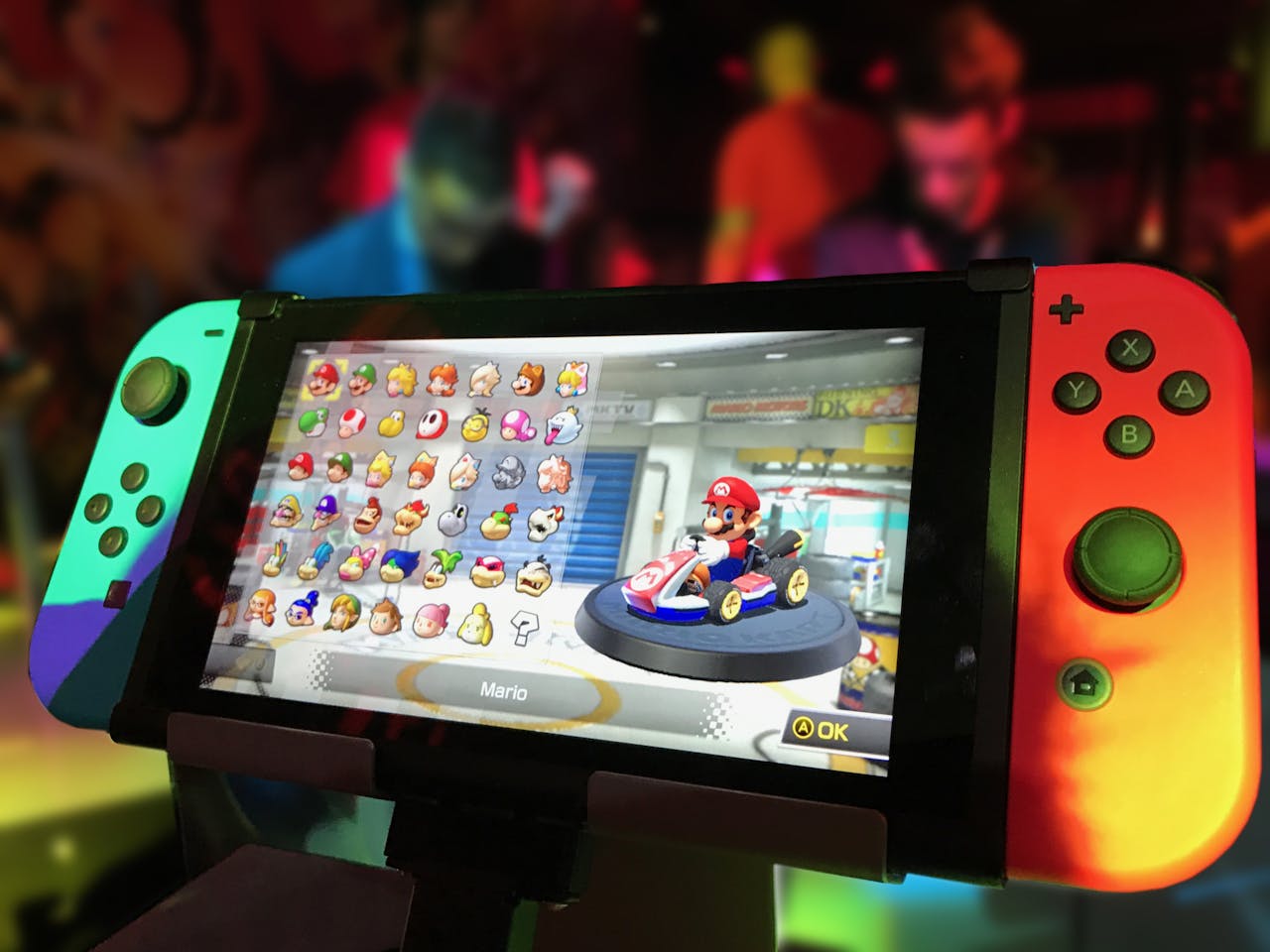 Services Close-up of a Nintendo Switch showing Mario Kart selection screen, gaming atmosphere in the background.